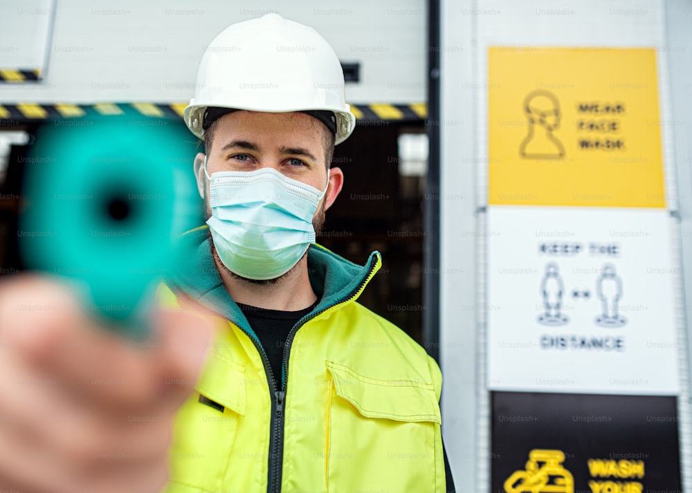 Portrait of worker with face mask in front of warehouse, coronavirus and temperature measuring concept.