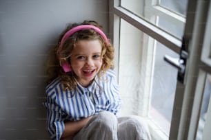 Porträt eines fröhlichen kleinen Mädchens mit Kopfhörern drinnen zu Hause, das auf der Fensterbank sitzt.