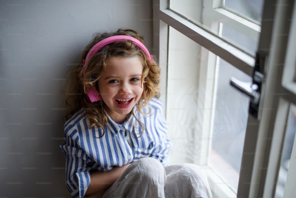 Portrait of cheerful small girl with headphones indoors at home, sitting on window sill.