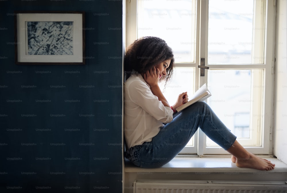 A portrait of young woman with book on window sill indoors at home, reading.