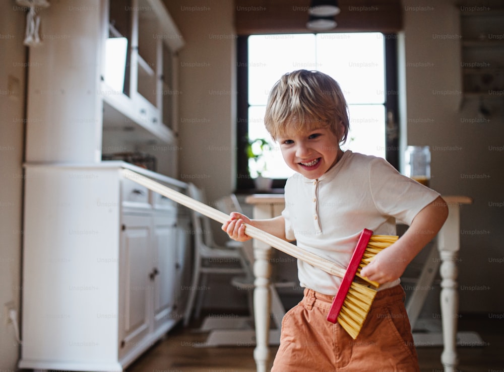 Um menino pequeno com vassoura ajudando com tarefas domésticas dentro de casa, se divertindo.