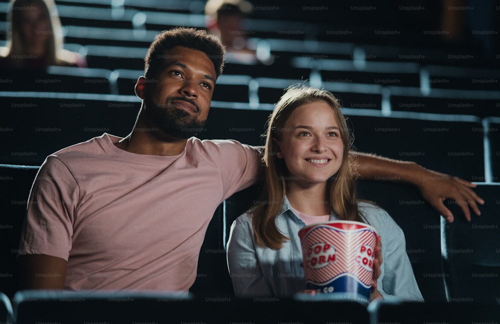 A front view of cheerful young couple with popcorn in the cinema, watching film.