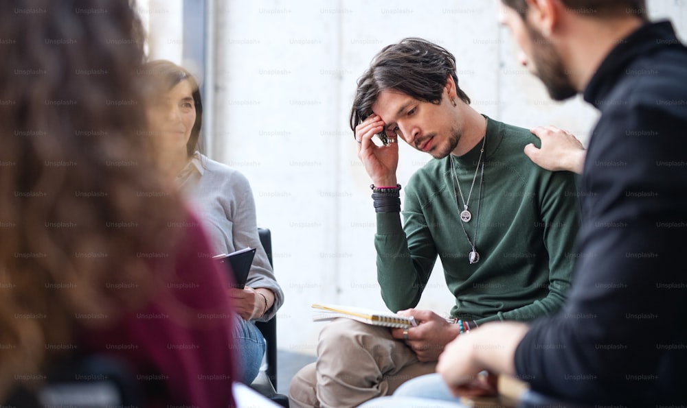 A sad and depressed man sitting in circle on group therapy, others supporting him.