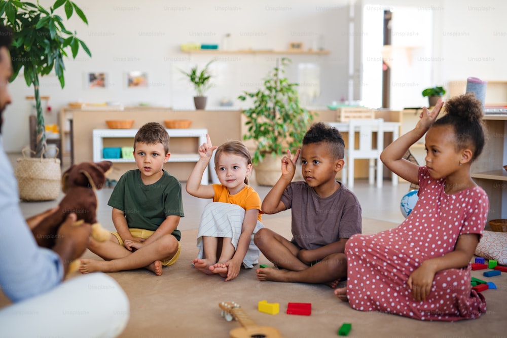 A group of small nursery school children with man teacher sitting on floor indoors in classroom, raising hands.