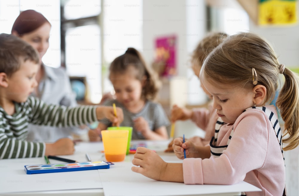 Un groupe de petits enfants de maternelle avec un enseignant à l’intérieur dans la salle de classe, en train de peindre.