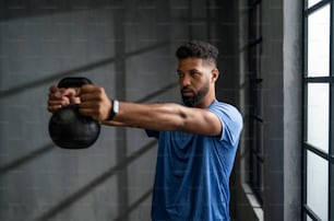 Um jovem esportista afro-americano de pé e levantando uma campainha de chaleira dentro de casa, conceito de treinamento.