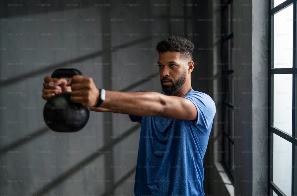 Ein junger afroamerikanischer Sportler steht und hebt eine Kettlebell in Innenräumen, Trainingskonzept.