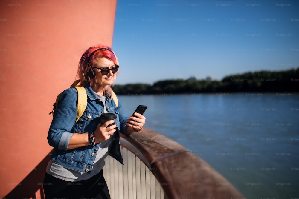 Senior woman with purple pink hair and smartphone and headphones standing outdoors in city or town.