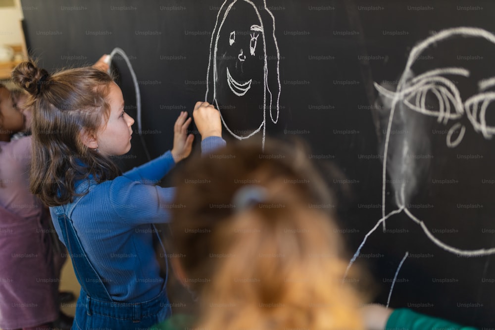 Little girls drawing with chalks on a blackboard wall indoors in playroom.