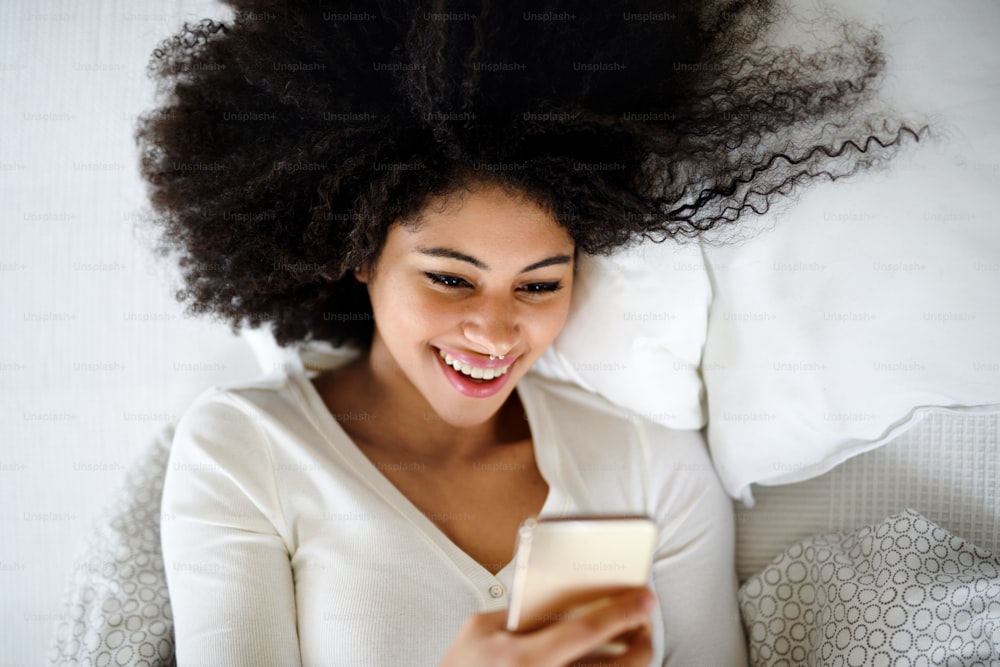 Top view portrait of happy young woman indoors on bed, using smartphone.