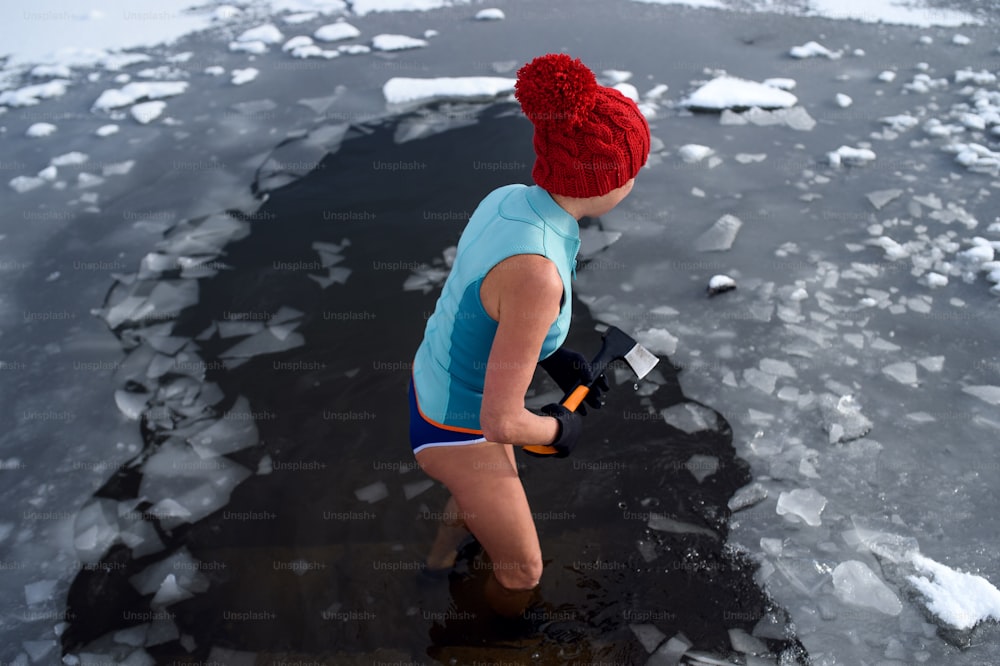 HIgh angle view of an active senior woman in swimsuit breaking ice with axe outdoors in winter, cold therapy concept.