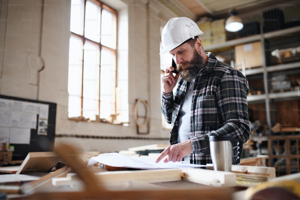 Un retrato de un carpintero masculino maduro mirando planos, planos y haciendo una llamada telefónica en el taller de carpintería. Concepto de pequeña empresa.