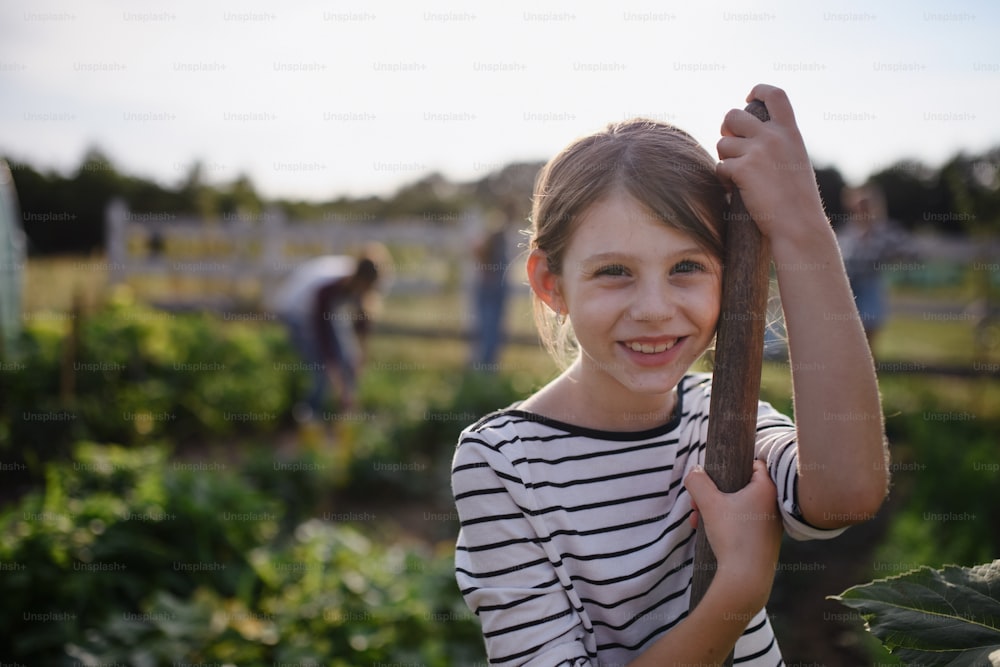 Porträt eines glücklichen kleinen Bauernmädchens mit Gartengerät draußen auf dem Gemeinschaftsbauernhof, mit Blick in die Kamera.