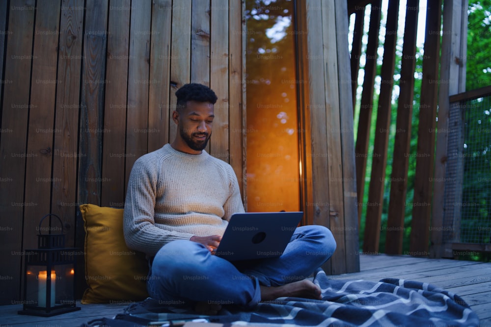 A happy young man with laptop resting outdoors in a tree house, weekend away and remote office concept.
