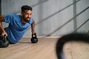 A young African American sportsman doing push ups with kettle bells indoors, workout training concept.