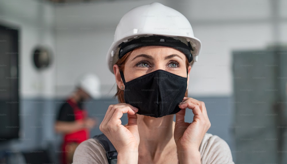 A portrait of woman worker with helmet indoors in factory putting on face mask.