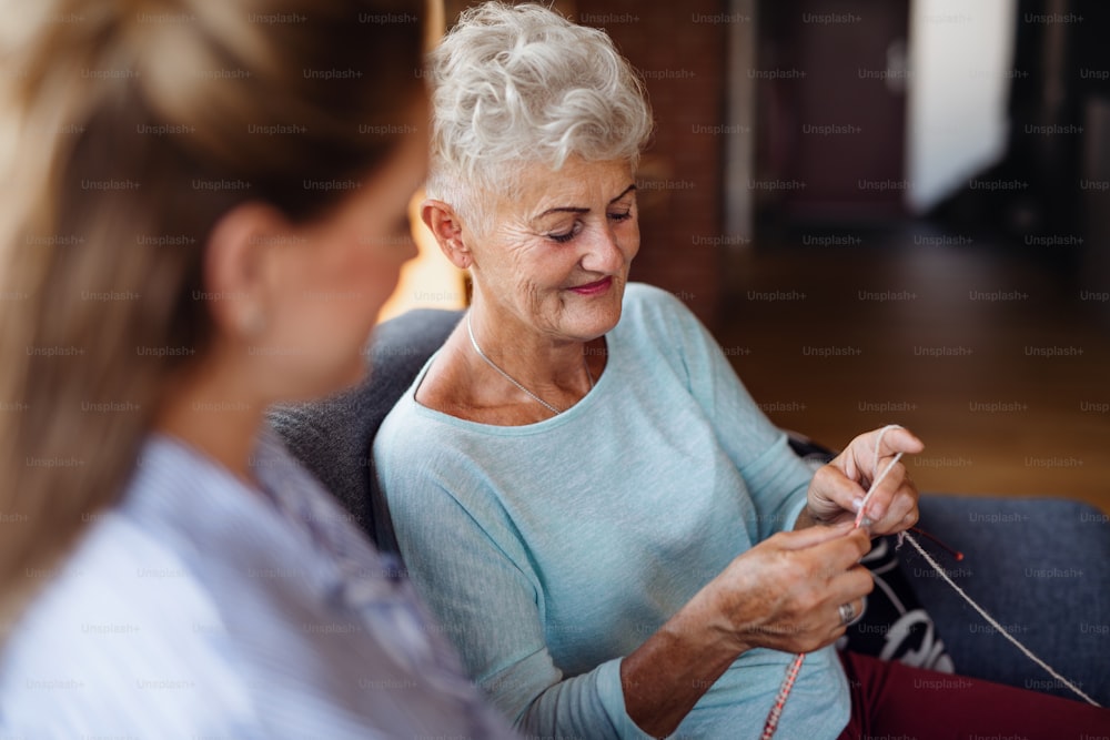 A happy senior woman sitting on sofa and learning to knit her adult daughter indoors at home.