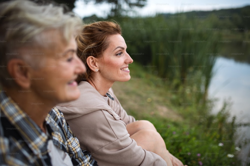 A profile view of happy senior mother hiker sitting with adult daughter by lake outdoors in nature