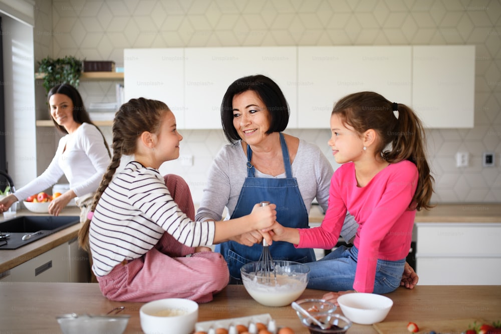 Bambine felici con la madre e una nonna che preparano la miscela di frittelle al chiuso a casa, cucinando.