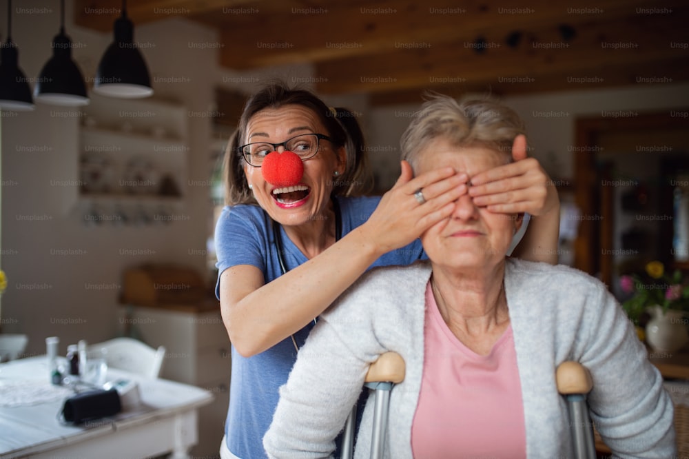 Healthcare worker or a caregiver with red nose visiting senior woman indoors at home, having fun and covering her eyes.