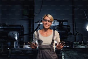 Portrait of a happy young industrial woman with dirty hands looking at camera indoors in metal workshop.