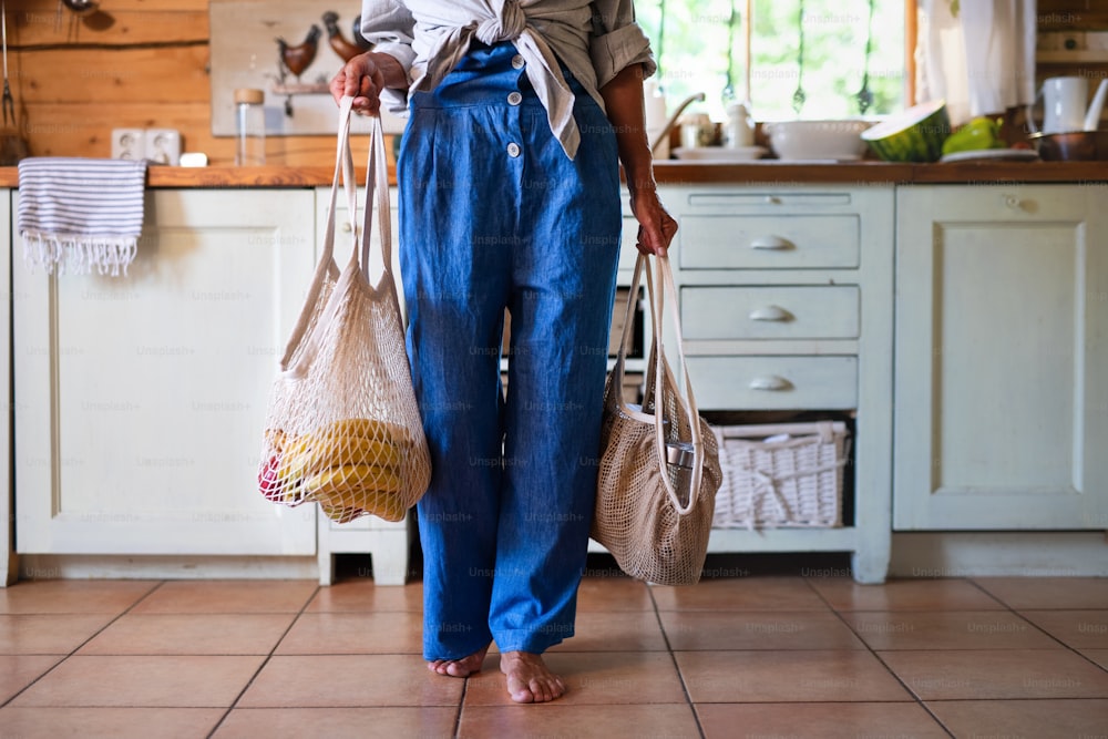 Une femme âgée tenant des sacs à provisions réutilisables à l’intérieur à la maison, un mode de vie durable.