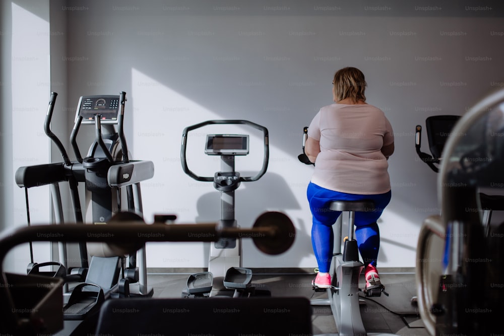 A rear view of mid adult overweight woman exercising on stationary bike indoors in gym