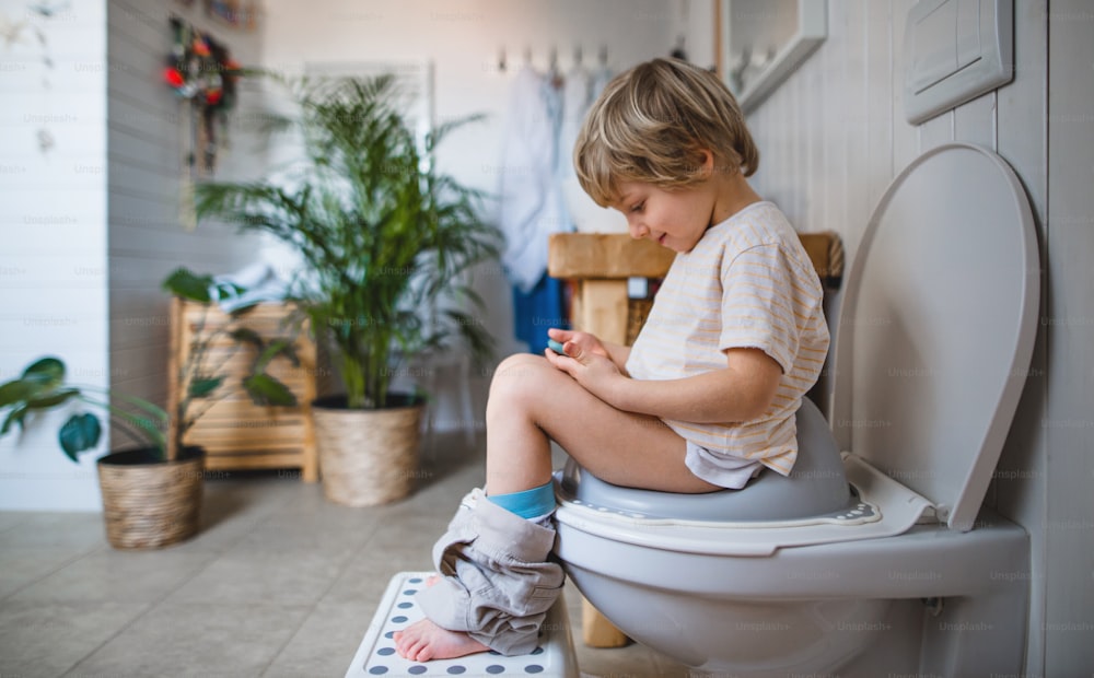 Vista lateral de un lindo niño pequeño sentado en el inodoro en el interior de la casa, usando un teléfono inteligente.