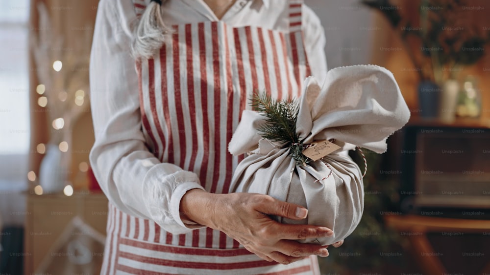 Un primer plano de una mujer mayor sosteniendo un regalo de Navidad envuelto en materiales naturales en el interior de casa.