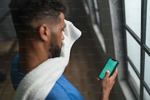 A high angle view of young African American sportsman standing indoors at gym, using smartphone.