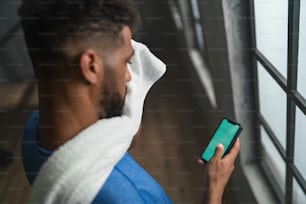 若いアフリカ系アメリカ人のスポーツマンがジムで屋内に立ち、スマートフォンを使用している高角度のビュー。