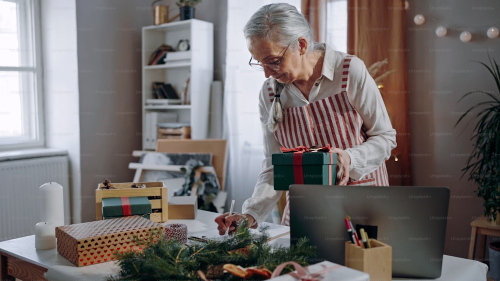 Una mujer mayor empacando regalos de Navidad y escribiendo notas para enumerar en el interior, concepto de pequeña empresa.