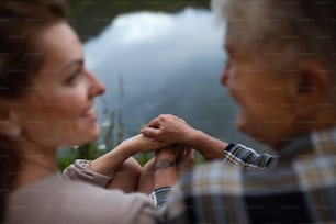 A rear view of senior mother holding hands with adult daughter sitting by lake in nature, focus on hands