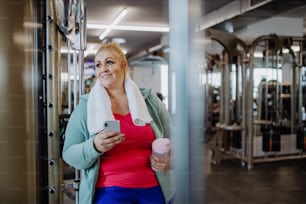 A happy mid adult overweight woman resting with mobile after exercise indoors in gym