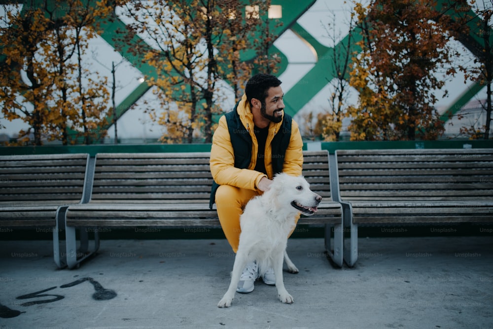 Um jovem feliz sentado no banco e segurando seu cachorro ao ar livre na cidade no outono.