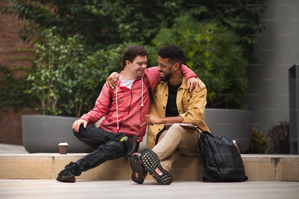 A happy young man with Down syndrome and mentoring friend sittingwith arms around outdoors