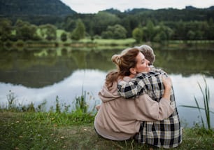 A rear view of happy senior mother embracing with adult daughter when sitting by lake outdoors in natureA close up of senior mother sitting on grass with adult daughter leaning her head on her outdoors.