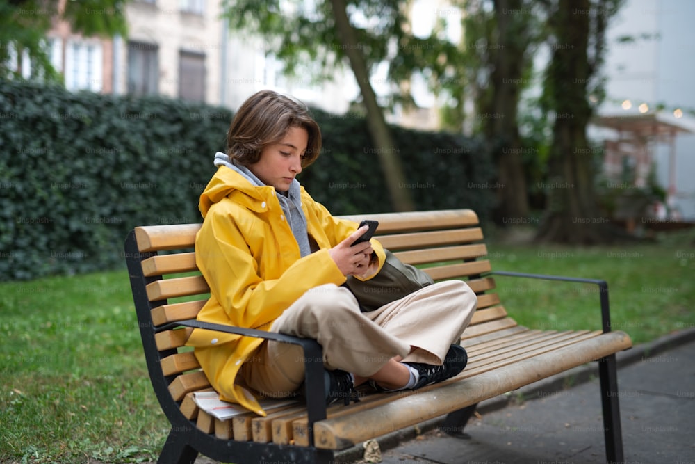 A preteen schoolgirl sitting on bench and using smartphone outdoors in town.
