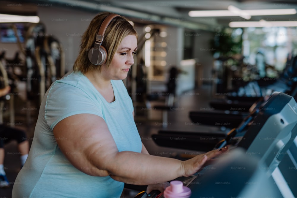 Mid adult overweight woman with headphones running on treadmill in gym