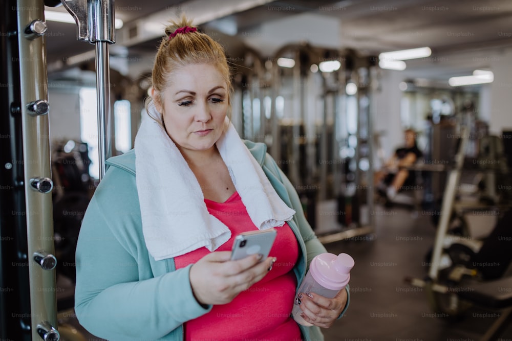 A happy mid adult overweight woman resting with mobile after exercise indoors in gym