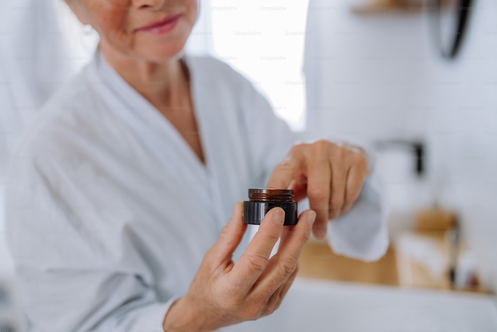 Una bella donna anziana in accappatoio che applica una crema naturale per il viso in bagno, nella cura della pelle e nel concetto di routine mattutina.