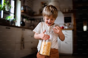 A portrait of cute small boy holding container with cornflakes indoors at home.