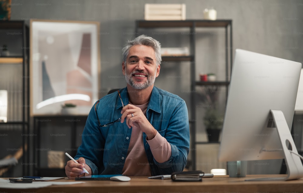 Un architecte d’homme mûr heureux travaillant sur une tablette à un bureau à l’intérieur d’un bureau, regardant la caméra.