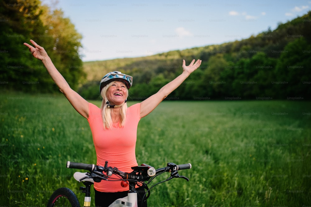 A cheerful active senior woman biker raising arms outdoors in nature.