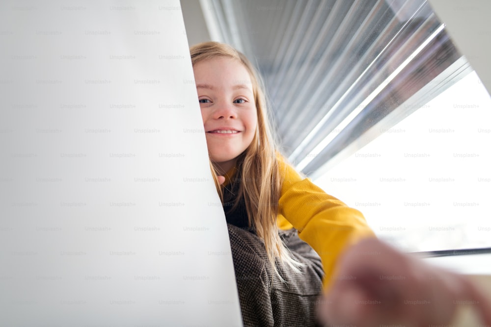A happy little girl with Down syndrome sitting on window at home.