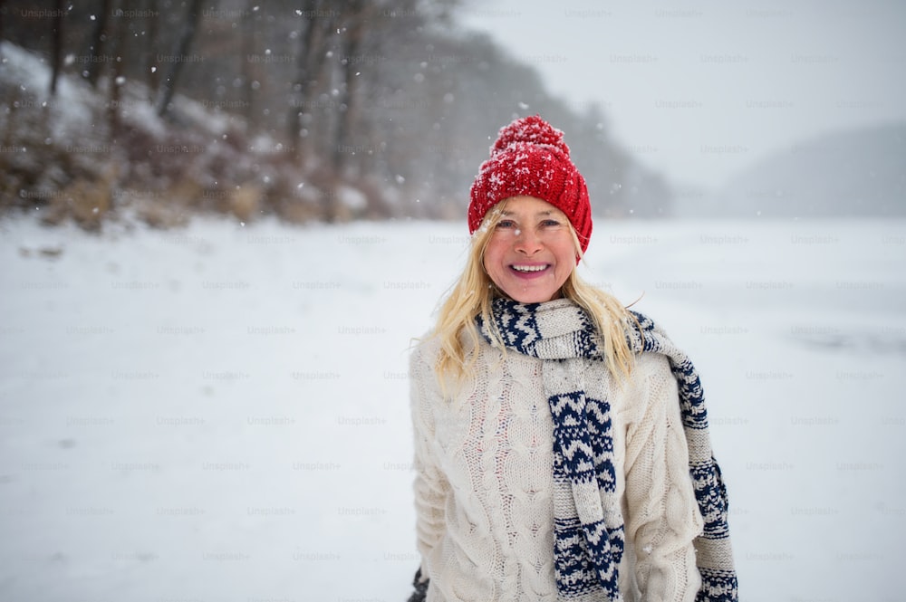 A front view portrait of happy senior woman with hat and outdoors standing in snowy nature, looking at camera.