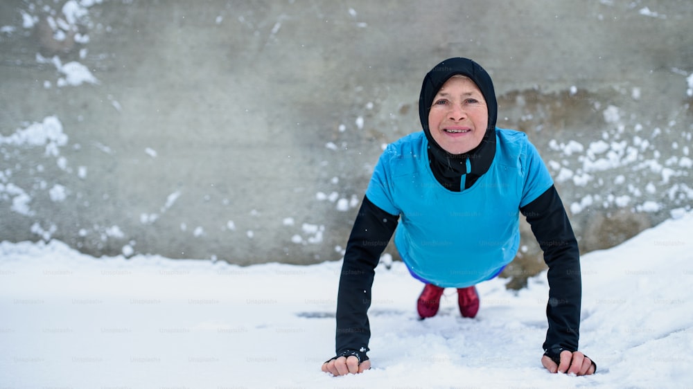 Front view of active senior woman outdoors in snowy winter, doing push ups.