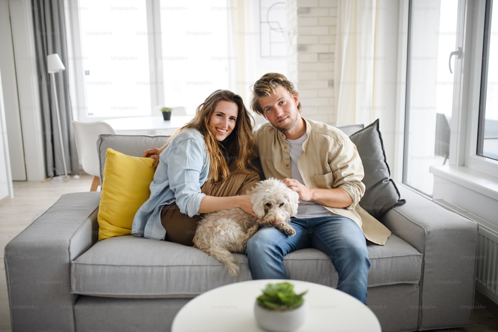 A happy young couple in love with dog sitting on sofa indoors at home, looking at camera.