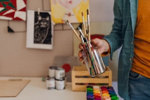 A close-up af art teacher's hand holding painbrushes indoors in classrom.