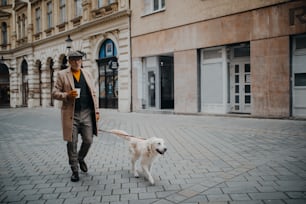 Ein eleganter älterer Mann mit Kaffee zum Mitnehmen geht im Winter mit seinem Hund draußen in der Stadt spazieren.